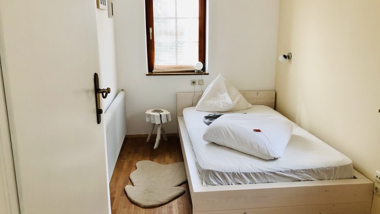 Ein kleines, gemütliches Schlafzimmer mit einem Einzelbett, Holzboden und einem kleinen Fenster mit Jalousie.