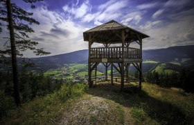Holzaussichtsturm mit Blick auf gr&uuml;ne T&auml;ler und Berge unter blauem Himmel.