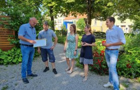 F&uuml;nf Personen stehen auf einem Kiesweg in einem Garten und unterhalten sich. Einer h&auml;lt ein Dokument in der Hand.