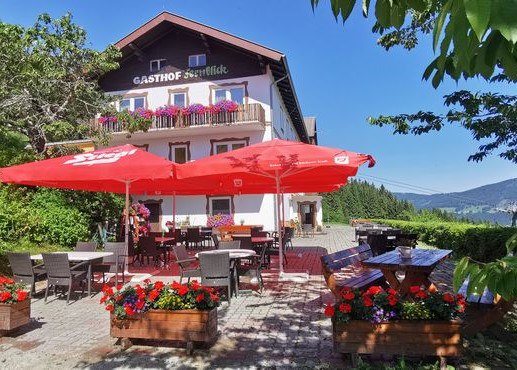 Ein traditioneller Gasthof mit Terrasse, roten Sonnenschirmen und Blumenkästen vor einem klaren blauen Himmel.