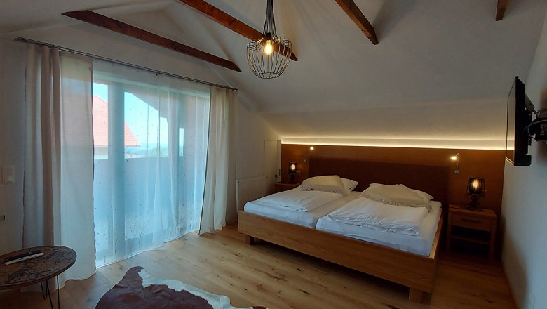 A cozy double room with a wooden floor, a large bed, curtains and a small table. A lamp hangs from the ceiling.
