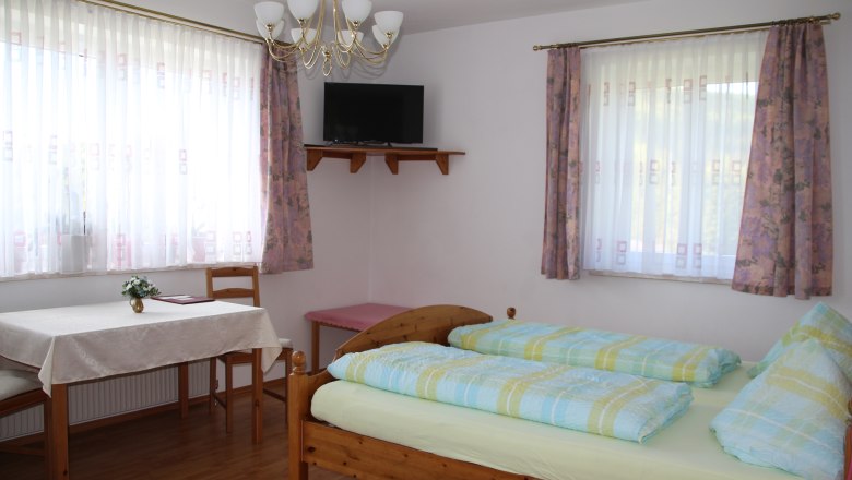 Ein helles Zimmer mit Doppelbett, Tisch, St&uuml;hlen und einem Fernseher an der Wand.