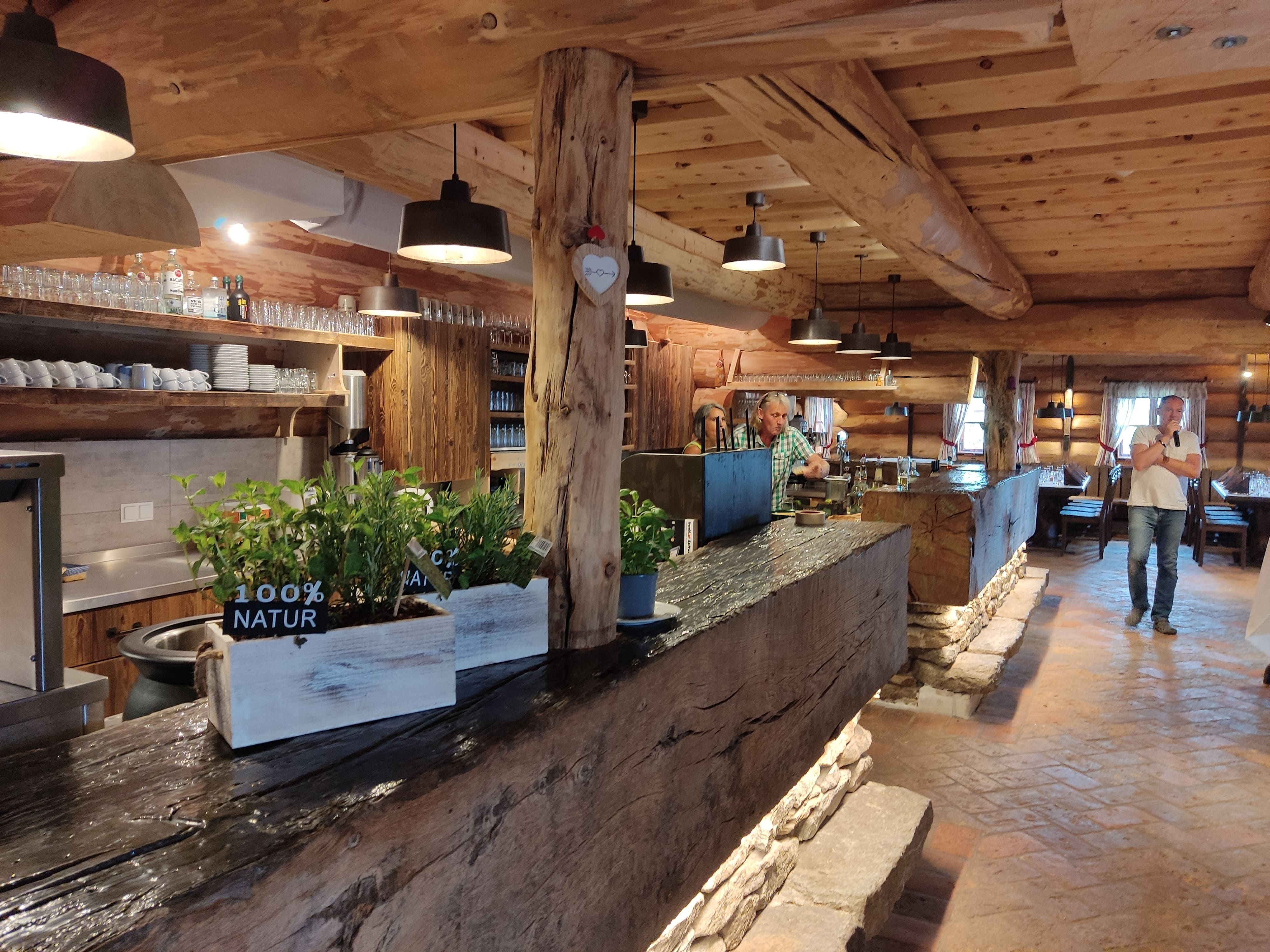 Innenansicht einer rustikalen Berghütte mit Holzbalken, Bar und Pflanzen.