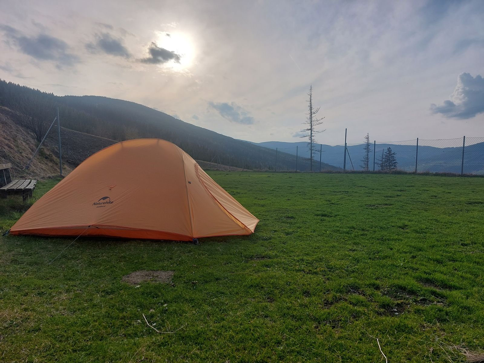 Ein orangefarbenes Zelt steht auf einer grünen Wiese, umgeben von Bergen und einem Zaun, bei bewölktem Himmel und Sonnenuntergang.
