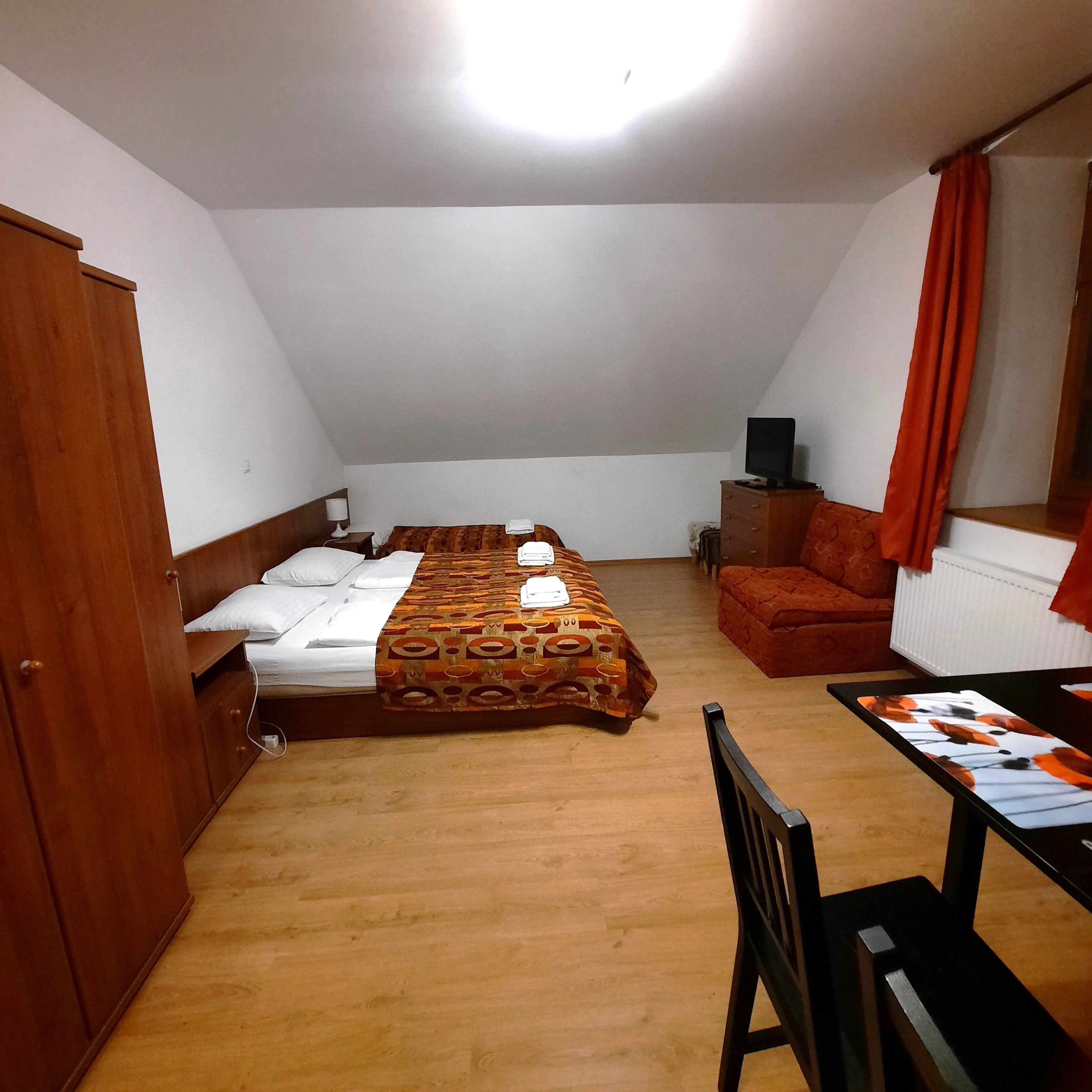 Ein einfach eingerichtetes Zimmer mit Doppelbett, Sofa, Tisch und Stuhl. Holzmöbel und orangefarbene Vorhänge dominieren den Raum.