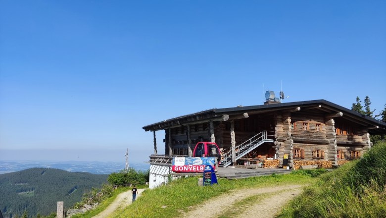 Eine Bergh&uuml;tte aus Holz mit einem roten Fahrzeug davor, umgeben von gr&uuml;nen Wiesen und einem klaren blauen Himmel.