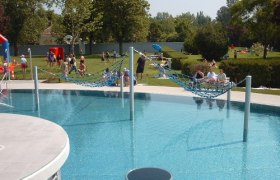 Ein Freibad mit H&auml;ngematten &uuml;ber dem Wasser und Menschen im Hintergrund auf der Liegewiese.