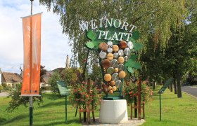 Willkommensschild in Platt mit Weintrauben-Dekoration und Kirche im Hintergrund.