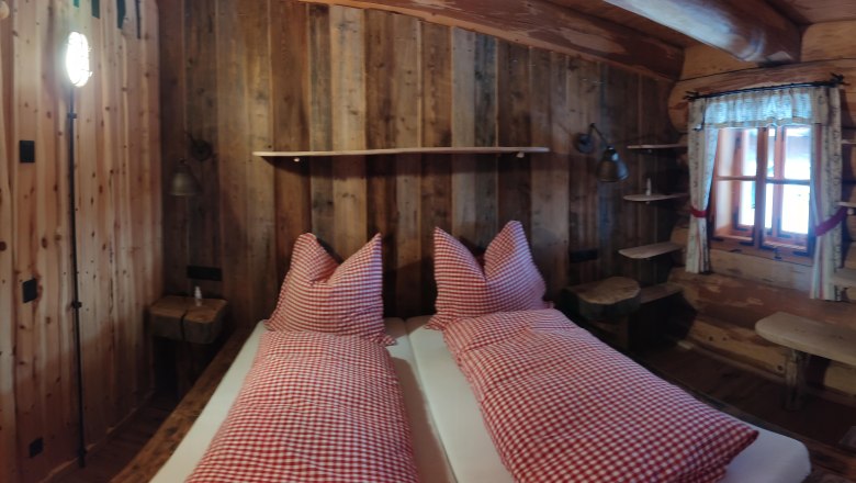 Rustikales Zimmer mit Holzw&auml;nden, Bett mit rot-wei&szlig; karierten Kissen, Fenster mit Vorh&auml;ngen, Holzt&uuml;r und Regalen.