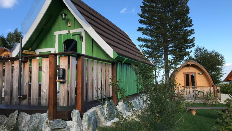 Glamping Park Mönichkirchen, © Glamping-Park Mönichkirchen OG Zwei kleine Holzhütten im Glamping-Stil mit grüner und brauner Fassade, umgeben von Bäumen und Gras.