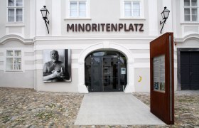 Eingang zum Forum Frohner am Minoritenplatz mit Kunstwerk und Infotafel.