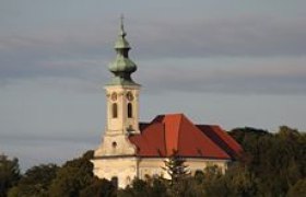 Pfarrkirche St. Nikolaus in Wolfpassing mit rotem Dach und gr&uuml;nem Turm vor bew&ouml;lktem Himmel.