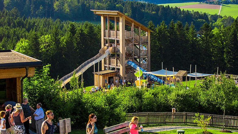 Rutschenturm aus Holz umgeben von Bäumen, und Menschen im Vordergrund.