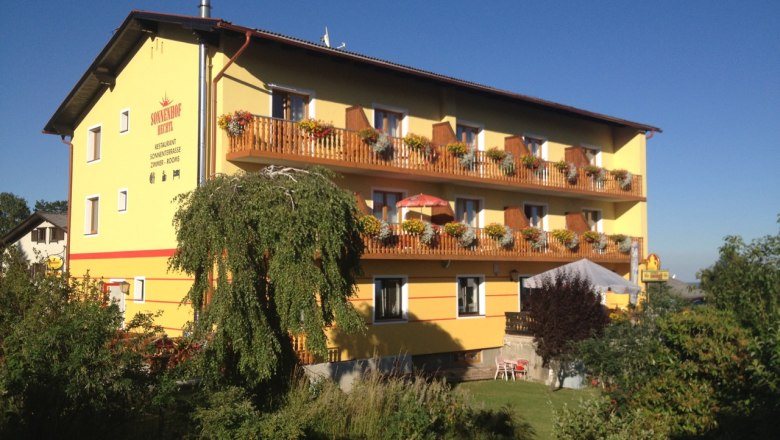 Sunny courtyard, © Johannes Hechtl Yellow building with wooden balconies and flowers, surrounded by trees and garden, under a blue sky.