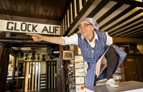 Eine Frau in traditioneller Kleidung zeigt in einem rustikalen Raum auf etwas. &Uuml;ber ihr steht "Gl&uuml;ck Auf".