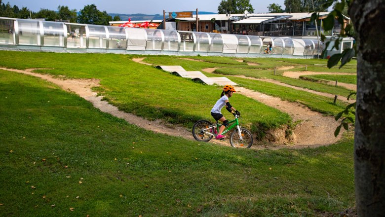 Ein Kind mit orangem Helm f&auml;hrt auf einem gr&uuml;nen Fahrrad durch einen Mini-Bikepark mit H&uuml;geln und Kurven.