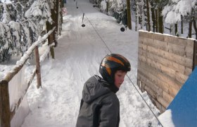 Ein Kind mit Helm steht auf Skiern an einem Schlepplift in einem verschneiten Wald.