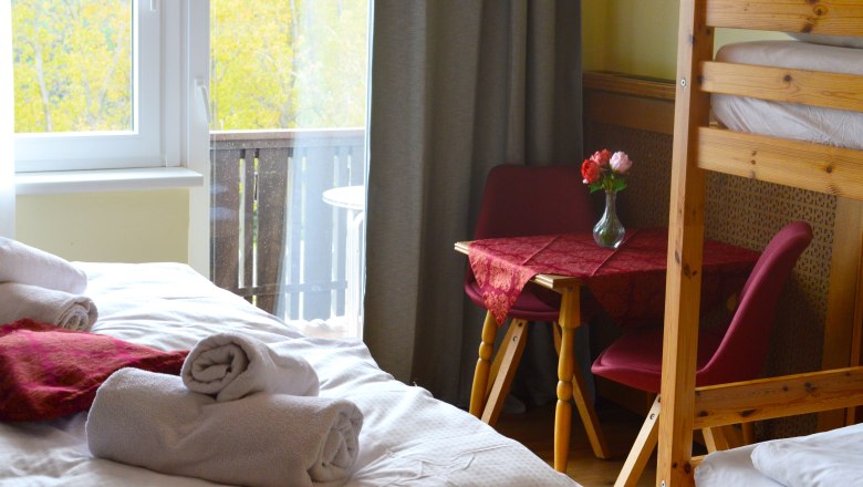 A cozy room with a bed, a table with red chairs and a bouquet of flowers. A window shows a balcony and autumnal trees.