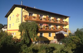 Gelbes Geb&auml;ude mit Holzbalkonen und Blumen, umgeben von B&auml;umen und Garten, unter blauem Himmel.
