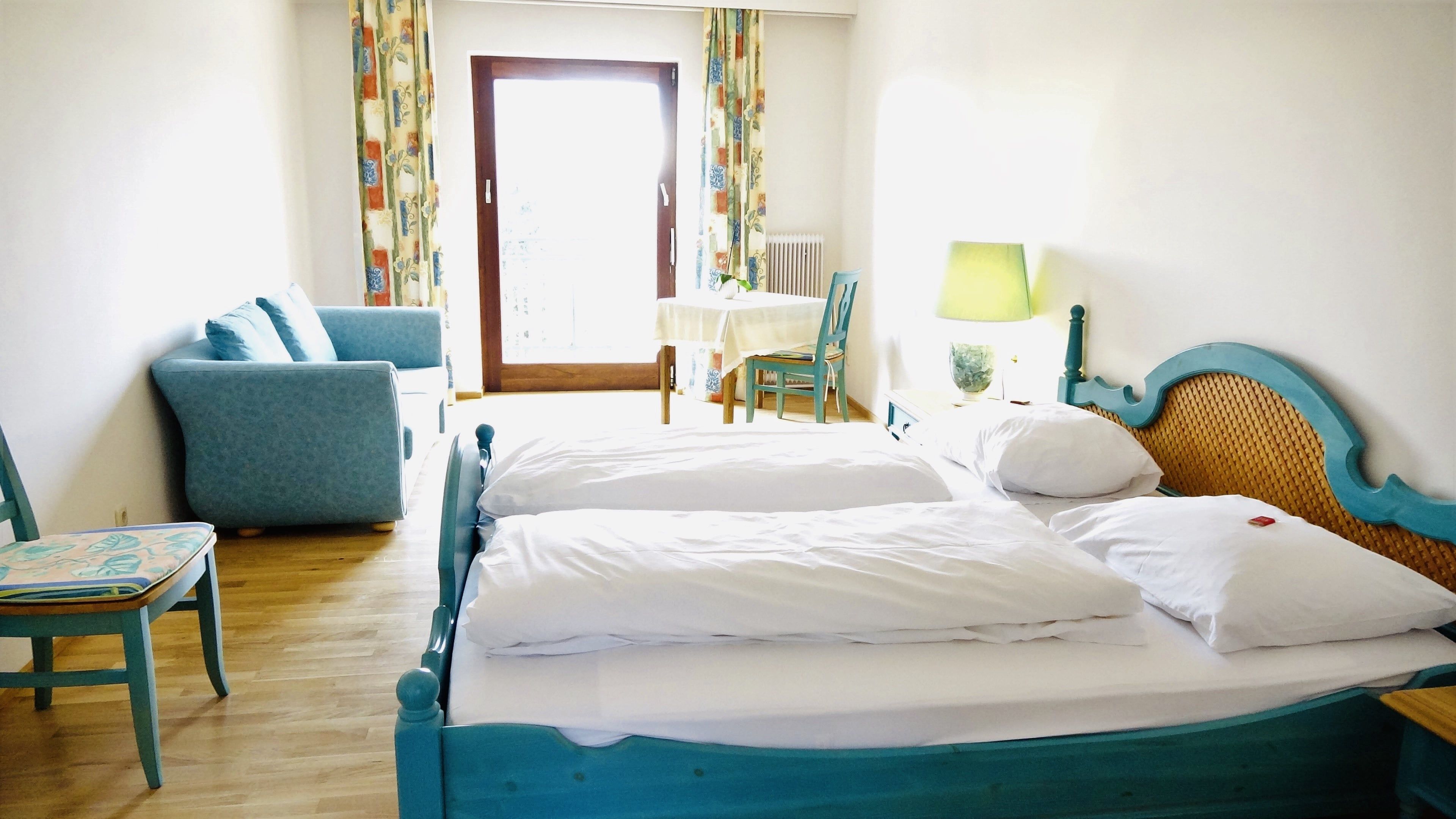Ein helles Zimmer mit einem Doppelbett, blauer Couch, Tisch und Stuhl vor einem Fenster mit Vorhängen.
