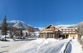 Winterlandschaft mit Hotel Forellenhof vor schneebedeckten Bergen und blauem Himmel.