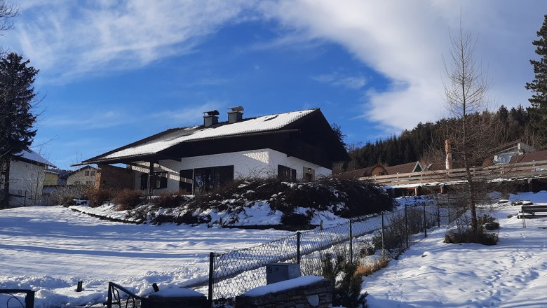 A snow-covered house with a dark roof and chimneys, surrounded by trees and a blue sky.