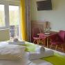 A cozy double room with bed, table, chairs and balcony. Yellow curtains and green accents create a friendly atmosphere.