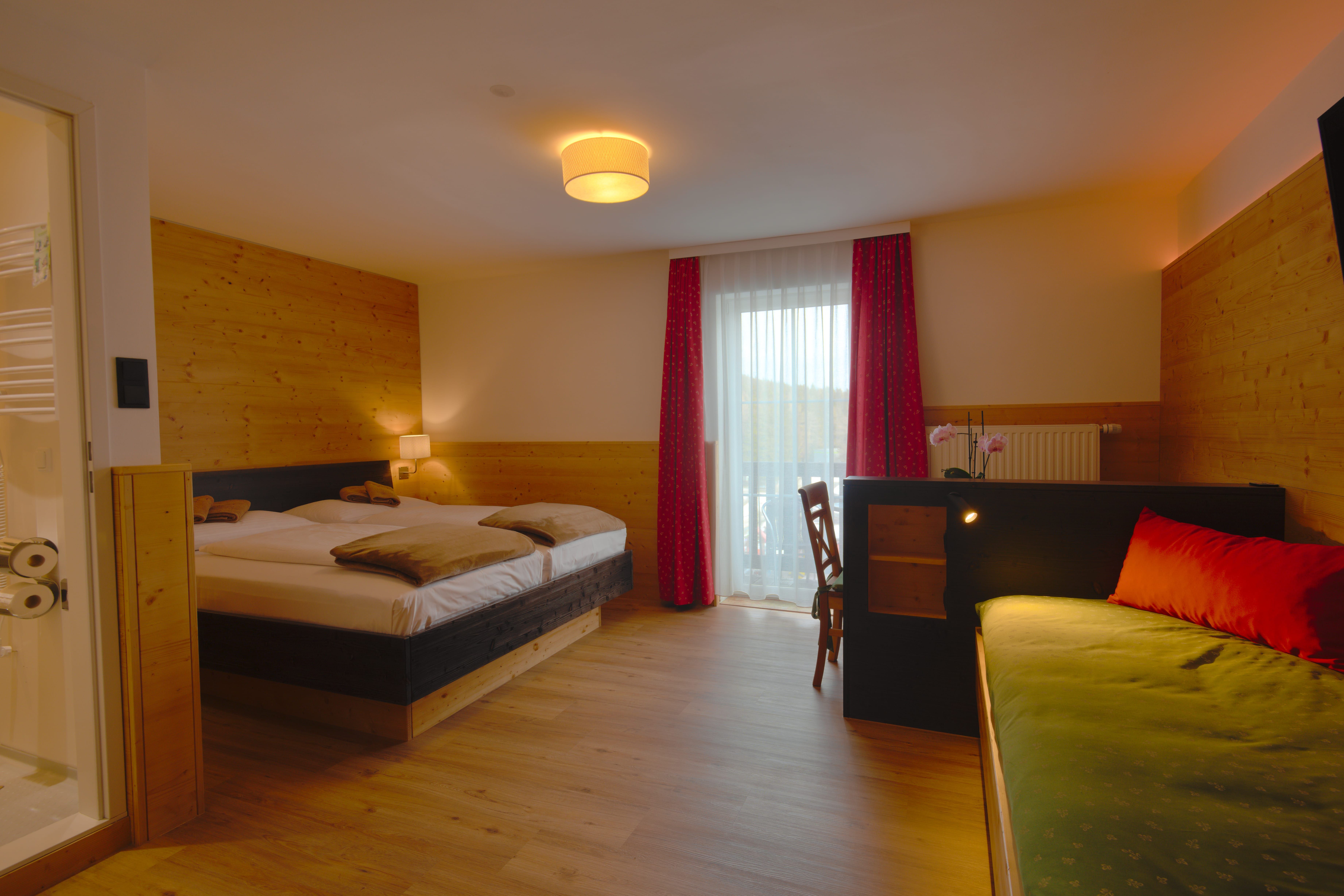 Ein gemütliches Doppelzimmer mit Holzwänden, einem Doppelbett, einem Sofa und einem Schreibtisch. Ein Fenster mit roten Vorhängen lässt Tageslicht herein.