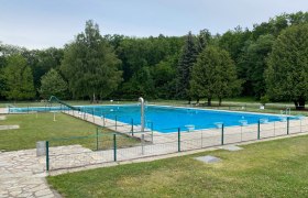 Ein leeres Freibad mit einem gro&szlig;en Schwimmbecken, umgeben von B&auml;umen und Rasenfl&auml;chen.