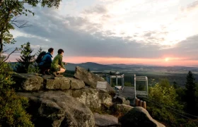 Zwei Personen auf einer Felsformation bei Sonnenuntergang mit Blick auf eine Aussichtsplattform und weite Landschaft.