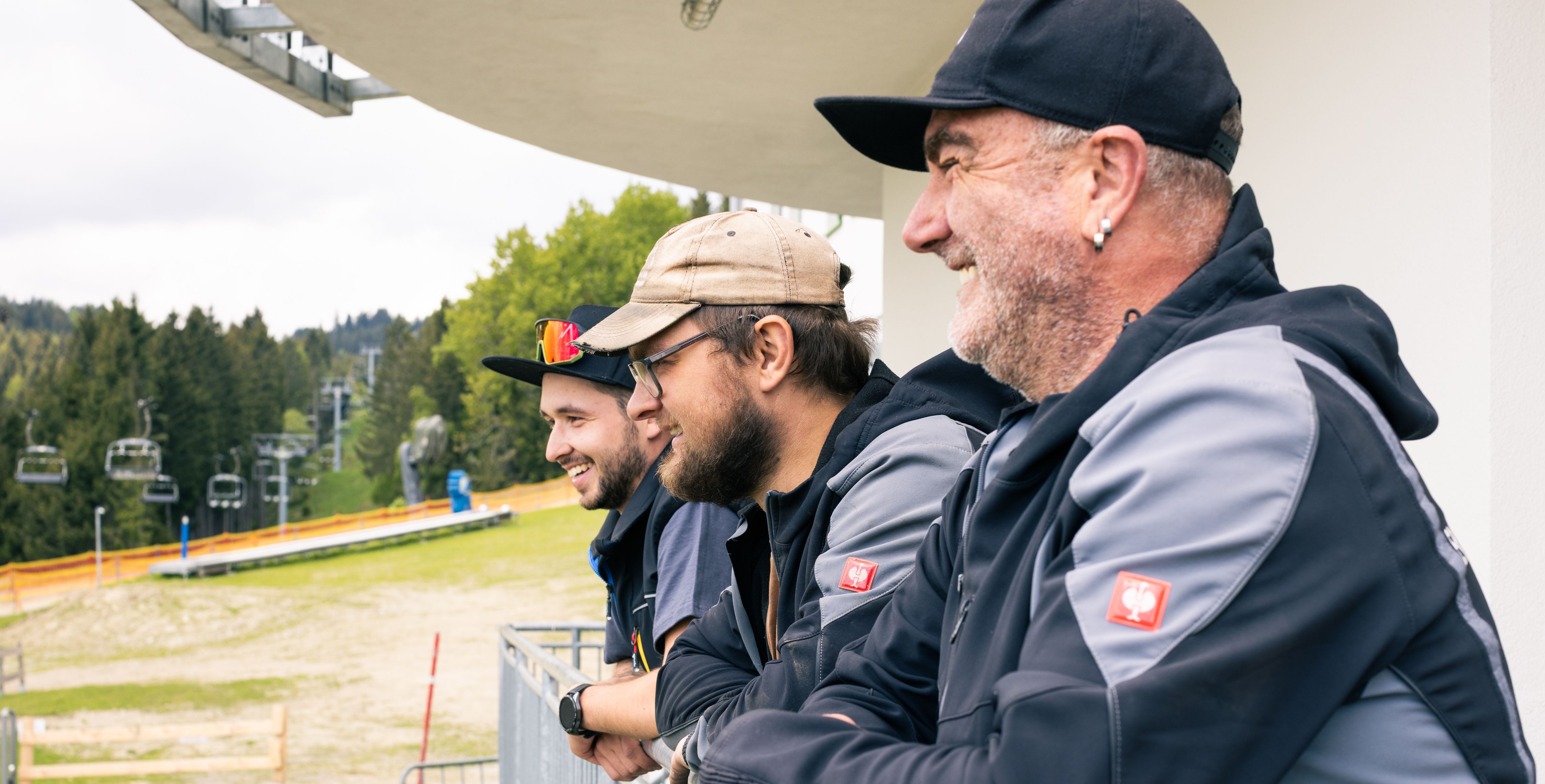 Drei Liftmitarbeiter stehen gemeinsam im Skigebiet und lachen freundlich in die Kamera. Sie tragen Arbeitskleidung und wirken gut gelaunt.