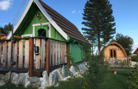 K&eacute;t kis fah&aacute;zik&oacute; glamping st&iacute;lusban, z&ouml;ld &eacute;s barna homlokzattal, f&aacute;kkal &eacute;s fűvel k&ouml;r&uuml;lv&eacute;ve.