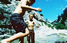 Zwei Kinder spielen im Wasser eines Gebirgsbaches, umgeben von Felsen und blauem Himmel.