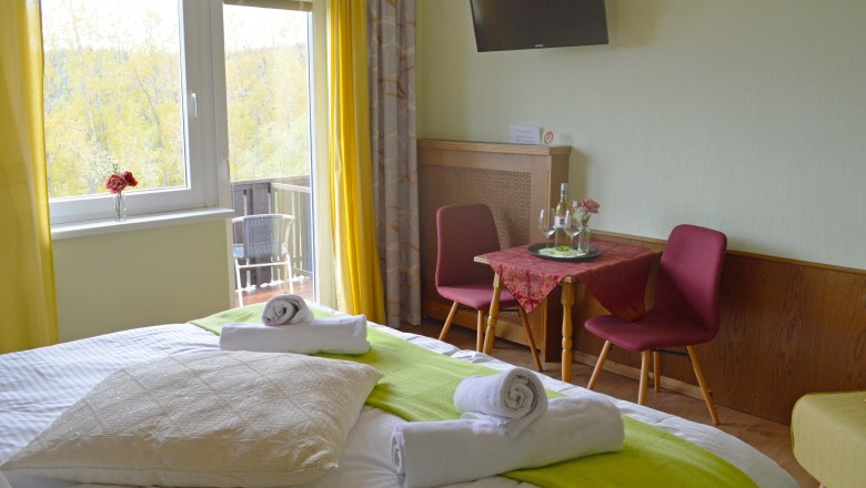 A cozy double room with bed, table, chairs and balcony. Yellow curtains and green accents create a friendly atmosphere.