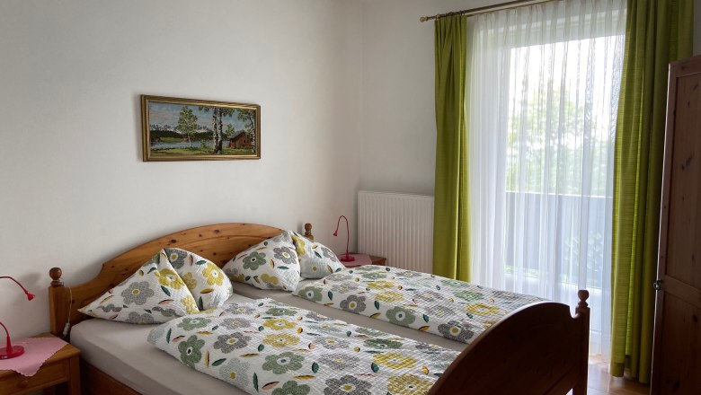 Ein gem&uuml;tliches Doppelzimmer mit Holzm&ouml;beln, einem gro&szlig;en Fenster mit gr&uuml;nen Vorh&auml;ngen und einem Landschaftsbild an der Wand.