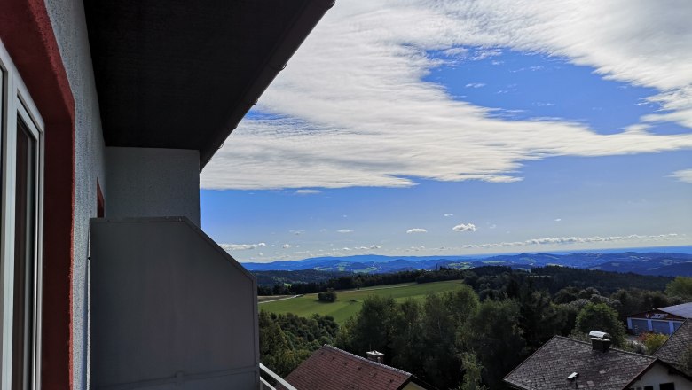 Blick von einem Balkon auf eine Landschaft mit Häusern, Bäumen und Hügeln unter einem blauen Himmel mit Wolken.