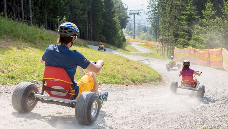 Personen fahren mit Mountaincarts einen Schotterweg hinunter, umgeben von Bäumen und einem Skilift.