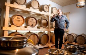 Ein Mann steht in einem Raum mit Holzf&auml;ssern, die mit 'Kraxados' und 'Whisky' beschriftet sind, und h&auml;lt ein Glas in der Hand.