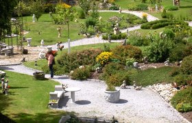 Ein gepflegter Schaugarten mit geschwungenen Wegen, Blumenbeeten und Gartenfiguren. Eine Person betrachtet die Pflanzen.