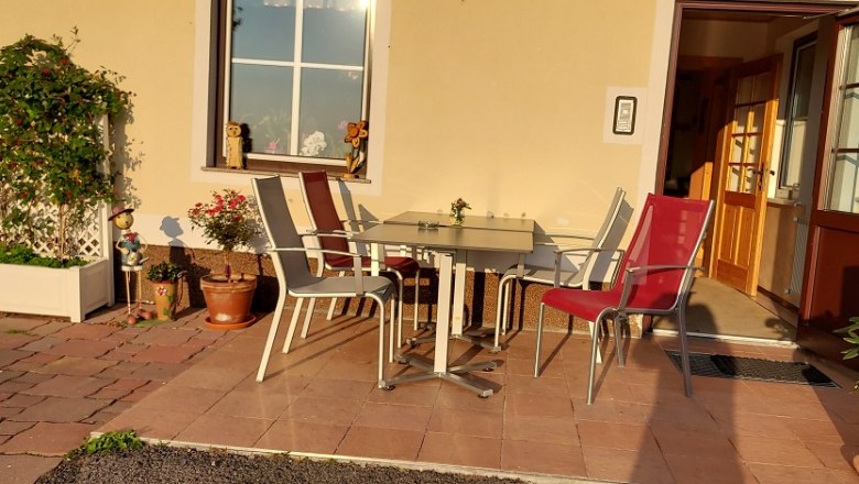 Terasz asztallal &eacute;s sz&eacute;kekkel egy &eacute;p&uuml;let előtt, n&ouml;v&eacute;nyekkel k&ouml;r&uuml;lv&eacute;ve.