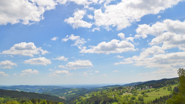 Panoramic view of a hilly landscape with forests and meadows under a blue sky with white clouds.